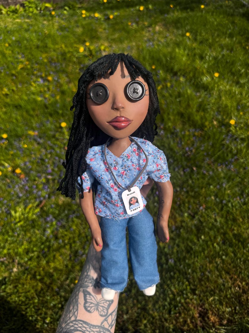 Peut inclure: Une poup&eacute;e &agrave; la peau fonc&eacute;e, aux cheveux en laine noire et aux yeux en boutons noirs. Elle porte un chemisier bleu clair &agrave; manches courtes avec un motif floral rose et blanc, un pantalon en jean bleu et des chaussures blanches. Un badge Brandi.