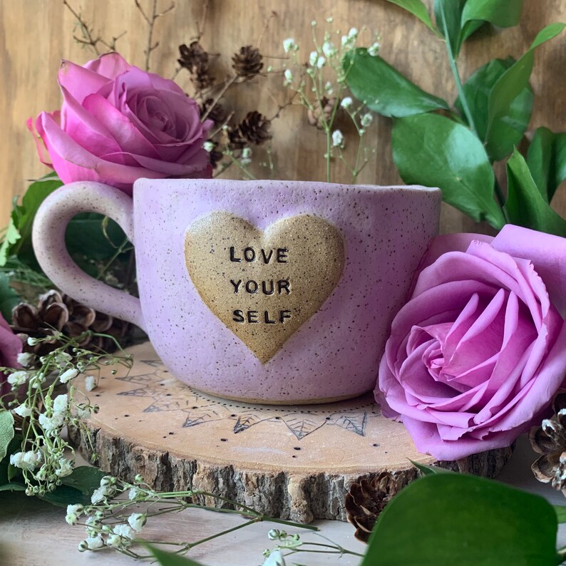 May include: A lavender ceramic mug with a gold heart that says "LOVE YOUR SELF" on it. The mug is sitting on a wooden coaster with a decorative design.