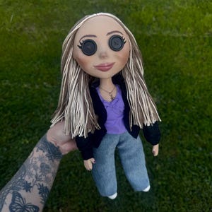 Peut inclure: Une poup&eacute;e aux longs cheveux raides en laine brune et blonde avec des yeux en boutons noirs. La poup&eacute;e porte un haut violet avec un petit n&oelig;ud, une veste noire et un pantalon en jean bleu. Un petit collier dor&eacute; avec un pendentif orne son cou.