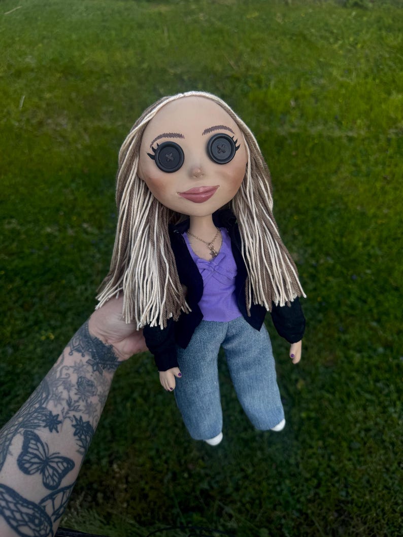 Peut inclure: Une poup&eacute;e aux longs cheveux raides en laine brune et blonde avec des yeux en boutons noirs. La poup&eacute;e porte un haut violet avec un petit n&oelig;ud, une veste noire et un pantalon en jean bleu. Un petit collier dor&eacute; avec un pendentif orne son cou.
