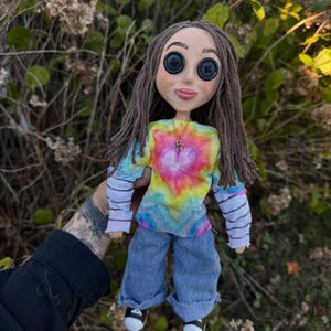 Peut inclure: Une poup&eacute;e aux cheveux en laine brune et aux yeux en boutons noirs, portant un t-shirt tie-dye &agrave; motif de c&oelig;ur, un haut blanc &agrave; manches longues avec des rayures horizontales noires, un jean bleu et des baskets noires et blanches avec un collier c&oelig;ur.