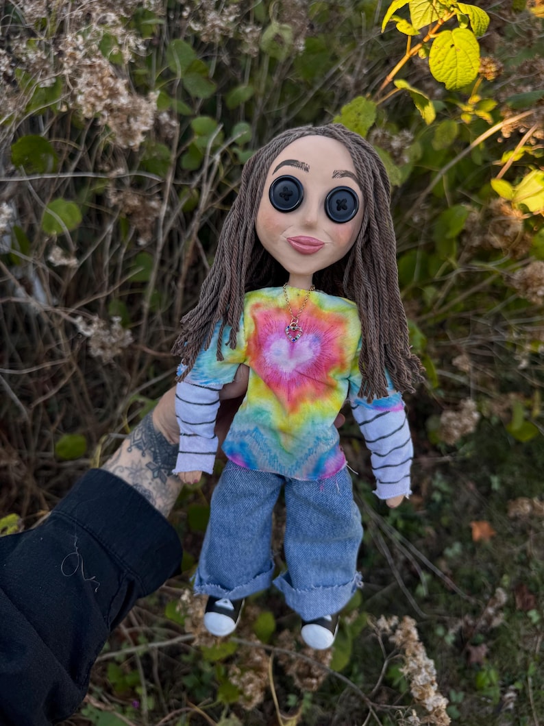 Peut inclure: Une poup&eacute;e aux cheveux en laine brune et aux yeux en boutons noirs, portant un t-shirt tie-dye &agrave; motif de c&oelig;ur, un haut blanc &agrave; manches longues avec des rayures horizontales noires, un jean bleu et des baskets noires et blanches avec un collier c&oelig;ur.