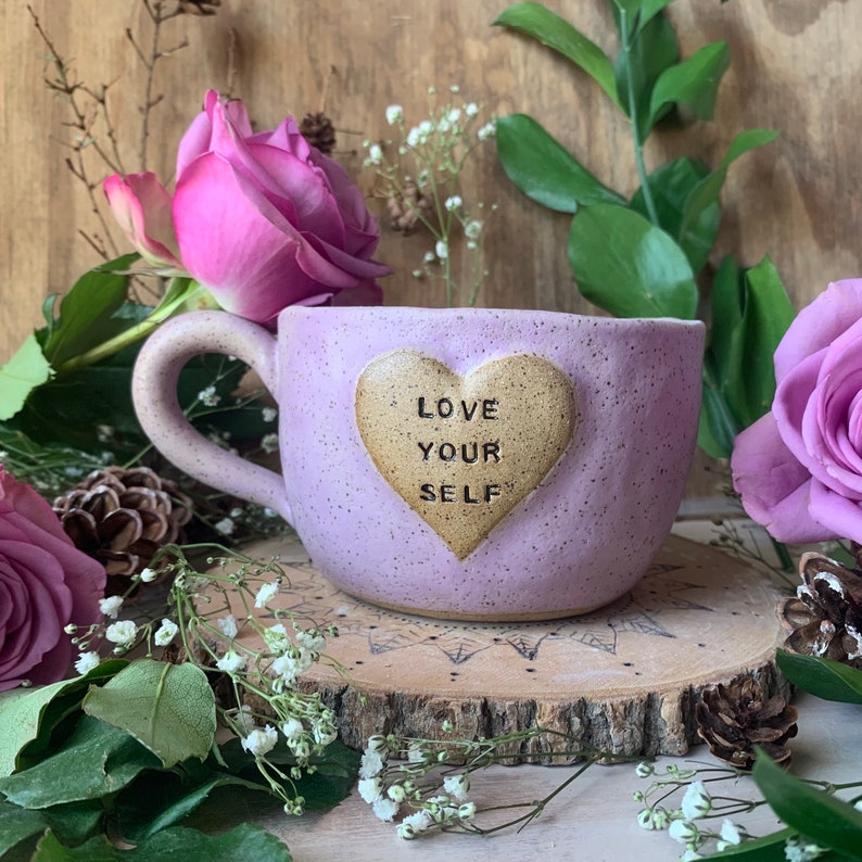May include: A lavender ceramic mug with a heart-shaped design that says "Love Your Self". The mug is sitting on a wooden surface with flowers and greenery around it.