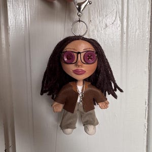 Peut inclure: Une poup&eacute;e porte-cl&eacute;s avec des cheveux en laine brun fonc&eacute;, des yeux en boutons violets et des lunettes &agrave; monture m&eacute;tallique noire. La poup&eacute;e porte une veste marron sur un haut blanc, un pantalon beige et des chaussures blanches, fix&eacute;e &agrave; un mousqueton.