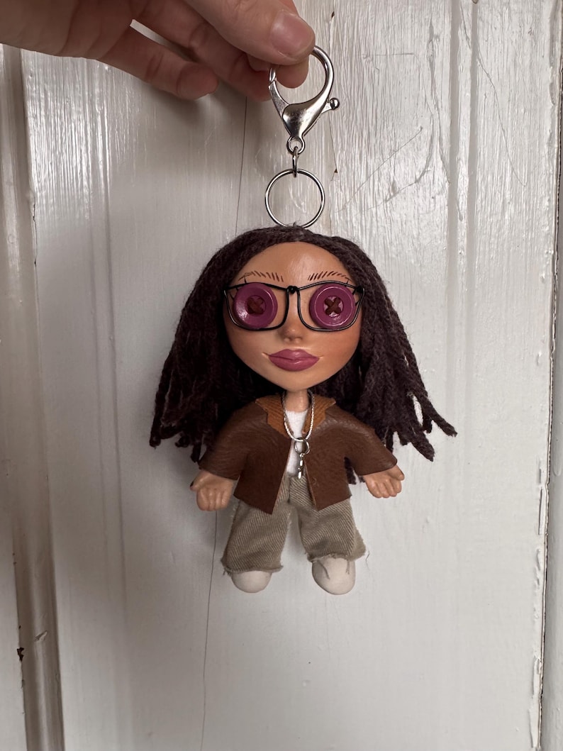 Peut inclure: Une poup&eacute;e porte-cl&eacute;s avec des cheveux en laine brun fonc&eacute;, des yeux en boutons violets et des lunettes &agrave; monture m&eacute;tallique noire. La poup&eacute;e porte une veste marron sur un haut blanc, un pantalon beige et des chaussures blanches, fix&eacute;e &agrave; un mousqueton.