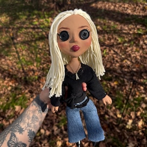 Peut inclure: Une poup&eacute;e aux longs cheveux en laine blonde, aux yeux en boutons noirs et aux traits du visage peints. La poup&eacute;e porte un haut noir &agrave; manches longues avec une encolure en dentelle, un collier dor&eacute; avec un pendentif lettre A et un pantalon en jean bleu.