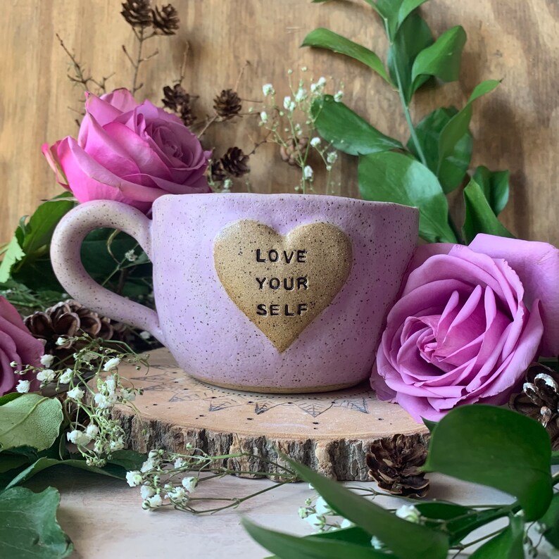 May include: Purple ceramic mug with a heart-shaped design that says "LOVE YOUR SELF" in gold lettering. The mug is sitting on a wooden surface with flowers.