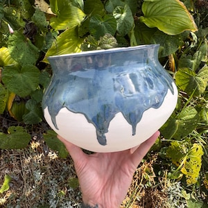 May include: A handmade ceramic bowl with a white base and a blue, dripped glaze. The bowl has a wide, open shape and is held in front of a background of green foliage. The blue glaze has a metallic sheen.