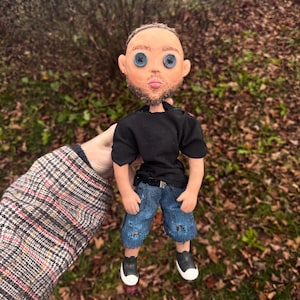 Peut inclure: Une poup&eacute;e faite &agrave; la main avec un teint clair, une courte barbe brune et des yeux en boutons bleus. La poup&eacute;e porte un t-shirt noir &agrave; manches courtes, un short en jean bleu avec des d&eacute;tails us&eacute;s et des baskets basses noires et blanches.