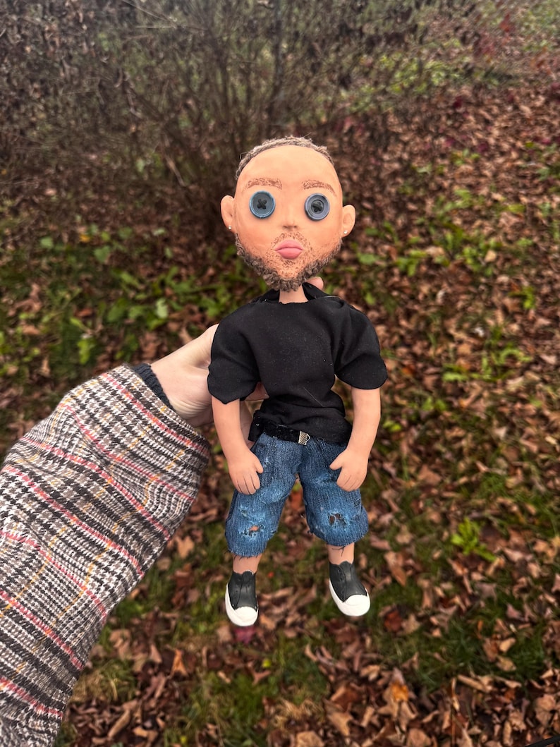 Peut inclure: Une poup&eacute;e faite &agrave; la main avec un teint clair, une courte barbe brune et des yeux en boutons bleus. La poup&eacute;e porte un t-shirt noir &agrave; manches courtes, un short en jean bleu avec des d&eacute;tails us&eacute;s et des baskets basses noires et blanches.