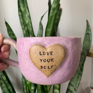 May include: A light purple ceramic mug with a brown heart-shaped accent. The heart has the words "LOVE YOUR SELF" inscribed on it.