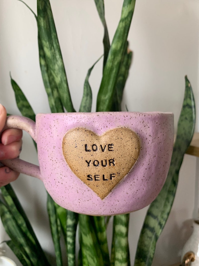 May include: A light purple ceramic mug with a brown heart-shaped accent. The heart has the words "LOVE YOUR SELF" inscribed on it.