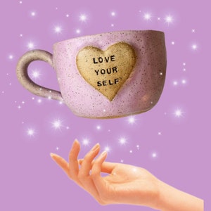 May include: A pink ceramic mug with a heart-shaped design that says "Love Your Self" in gold lettering. The mug is floating above a hand with sparkling lights in the background.