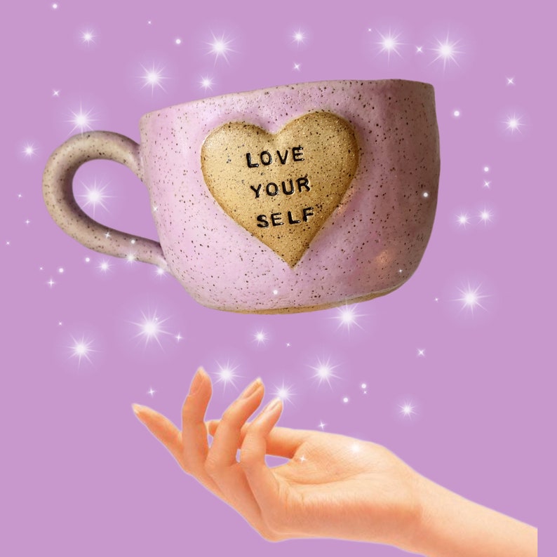 May include: A pink ceramic mug with a heart-shaped design that says "Love Your Self" in gold lettering. The mug is floating above a hand with sparkling lights in the background.