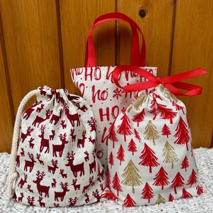 May include: Three fabric gift bags with festive patterns. One bag features red reindeer on a white background with a white drawstring. Another has red 'HO HO' text and red handles. The third has red and gold glitter trees with a red ribbon tie.