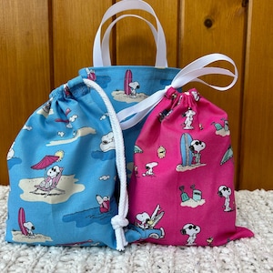 Op de afbeelding: Twee trekkoordtassen met een cartoon-hondendruk. De blauwe tas heeft een wit en blauwe print met een cartoon-hond die surft. De roze tas heeft een wit en roze print met een cartoon-hond die surft. Beide tassen hebben witte trekkoorden.