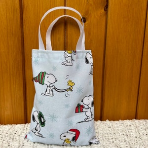 May include: A blue and white tote bag with a Christmas theme featuring Snoopy and Woodstock. The bag has white handles and a variety of Snoopy characters in different poses.