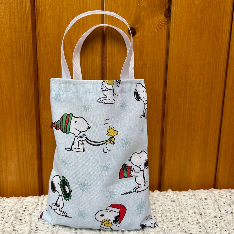 May include: A blue and white tote bag with a Christmas theme featuring Snoopy and Woodstock. The bag has white handles and a variety of Snoopy characters in different poses.
