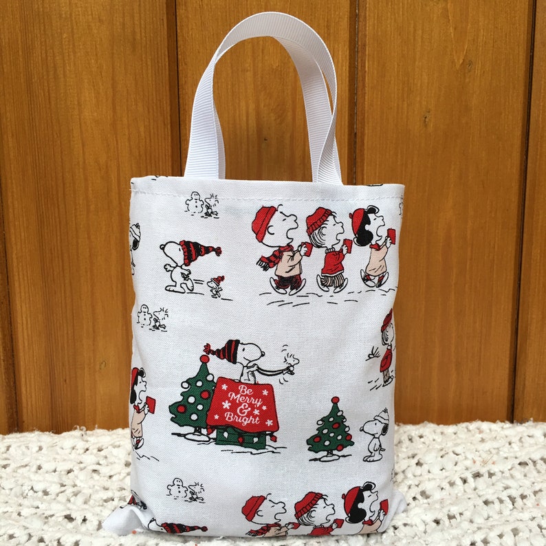 May include: White canvas tote bag with a Christmas theme featuring Peanuts characters. The bag has red and black accents and the text "Be Merry & Bright".