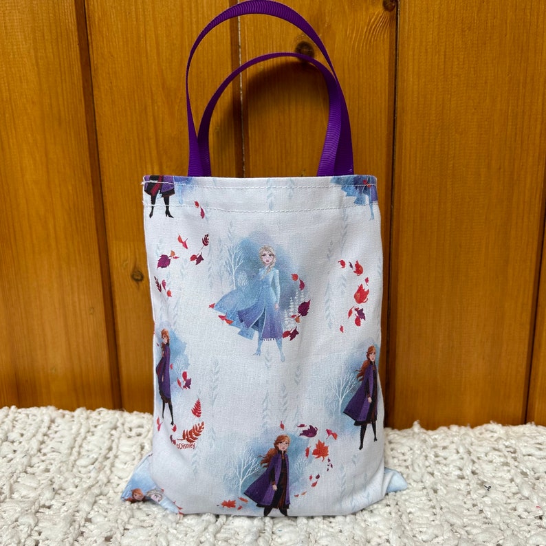 K&ouml;nnte beinhalten: Eine wei&szlig;e Tragetasche mit einem violetten Griff. Die Tasche zeigt ein sich wiederholendes Muster von Frozen-Charakteren, darunter Elsa, Anna und Olaf, vor einem hellblauen Hintergrund mit roten und wei&szlig;en Bl&auml;ttern.