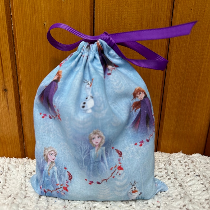 K&ouml;nnte beinhalten: Eine blaue Kordeltasche mit einem violetten Band. Die Tasche hat ein sich wiederholendes Muster mit Frozen-Charakteren, darunter Elsa, Anna, Olaf und Kristoff, auf hellblauem Hintergrund.