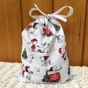 May include: White fabric gift bag with a drawstring closure. The bag features a repeating pattern of Peanuts characters celebrating Christmas. The characters include Snoopy, Charlie Brown, Lucy, and Linus. The bag has a white ribbon tied around the top. The bag has the text "Be Merry & Bright" printed on it.