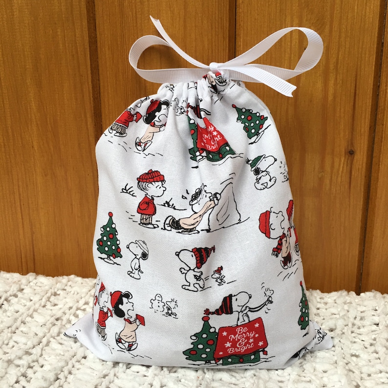 May include: White fabric gift bag with a drawstring closure. The bag features a repeating pattern of Peanuts characters celebrating Christmas. The characters include Snoopy, Charlie Brown, Lucy, and Linus. The bag has a white ribbon tied around the top. The bag has the text "Be Merry & Bright" printed on it.