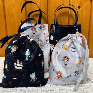 May include: Three fabric bags with a nautical theme. The bags are black and white with blue trim. The bags feature a variety of designs, including a ship, a pirate, and a cat. All bags are made of cotton and have a drawstring closure.