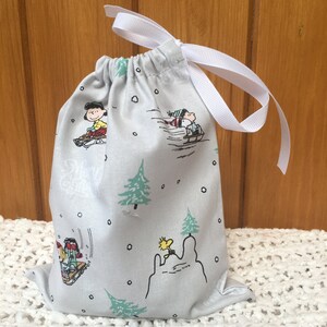 May include: A white drawstring bag with a white ribbon. The bag is decorated with a winter scene featuring the Peanuts characters, including Snoopy and Woodstock. The bag is perfect for gift giving or storing small items.