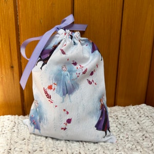 K&ouml;nnte beinhalten: Eine wei&szlig;e Kordeltasche mit einem lila Band. Die Tasche hat ein sich wiederholendes Muster mit Frozen-Charakteren, darunter Elsa und Anna, auf wei&szlig;em Hintergrund mit roten und violetten Akzenten.