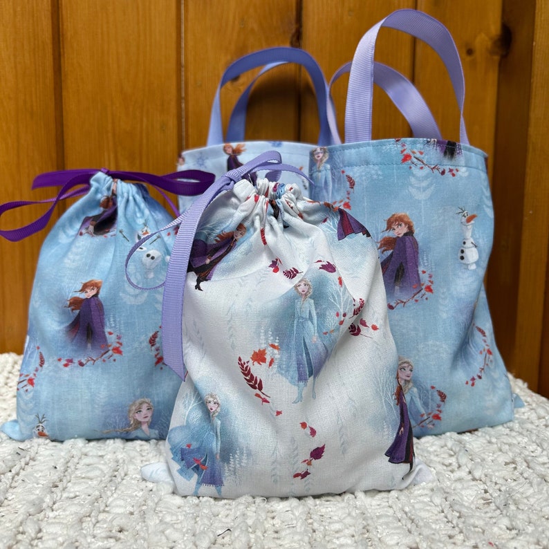 K&ouml;nnte beinhalten: Drei blaue Stofftaschen mit Frozen-Motiv. Die Taschen haben einen wei&szlig;en Hintergrund mit Bildern von Elsa, Anna und Olaf. Die Taschen haben violette Schleifenb&auml;nder.