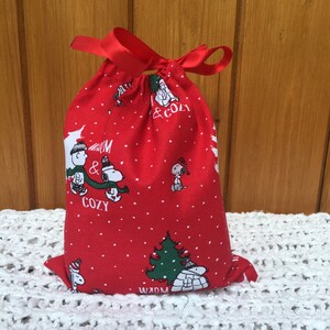 May include: Red drawstring bag with a white polka dot pattern. The bag features a Christmas-themed print of Snoopy and Woodstock with the text "Warm & Cozy".