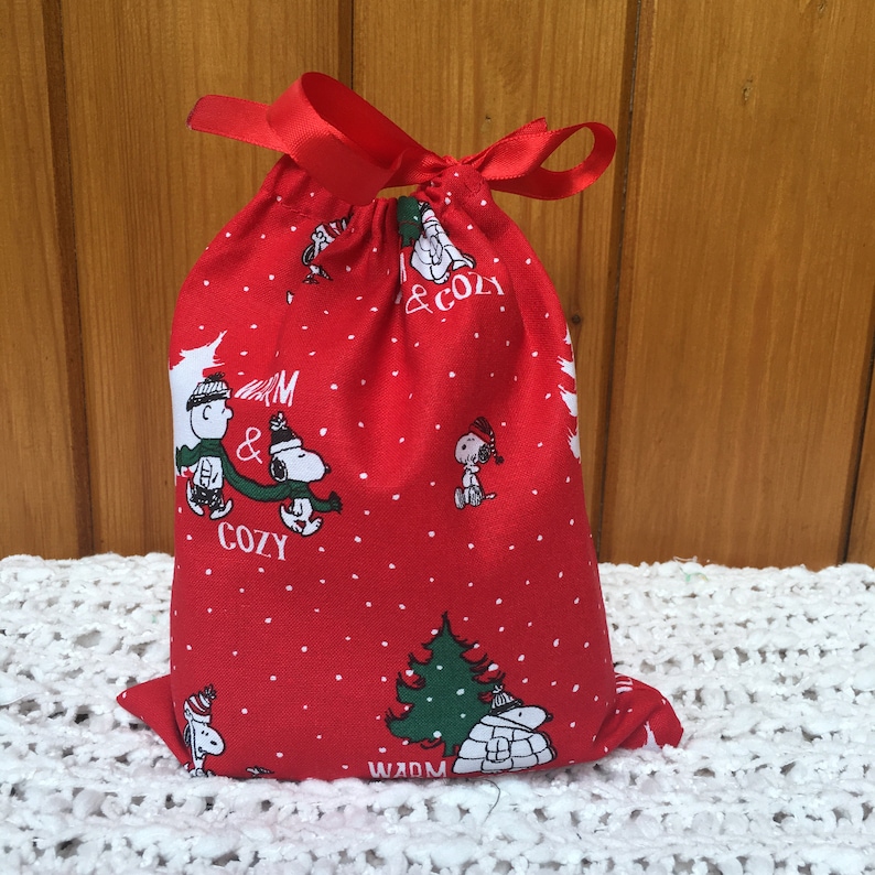 May include: Red drawstring bag with a white polka dot pattern. The bag features a Christmas-themed print of Snoopy and Woodstock with the text "Warm & Cozy".