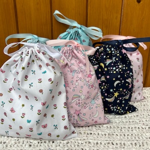 Könnte beinhalten: Vier Stoffbeutel mit Kordelzug und verschiedenen Mustern. Ein Beutel ist weiß mit kleinen rosa und blauen Blumen, ein anderer ist rosa mit weißen Sternen und Sternschnuppen, ein weiterer ist blau mit weißen Sternen und Sternschnuppen, und der letzte ist weiß mit rosa Blumen und einem blau-rosa Streifen.