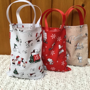 May include: Three small fabric gift bags with Christmas-themed Peanuts characters. The bags are white with red and black accents, red with white snowflakes and black accents, and beige with brown and black accents. The bags have white, red, and brown handles.