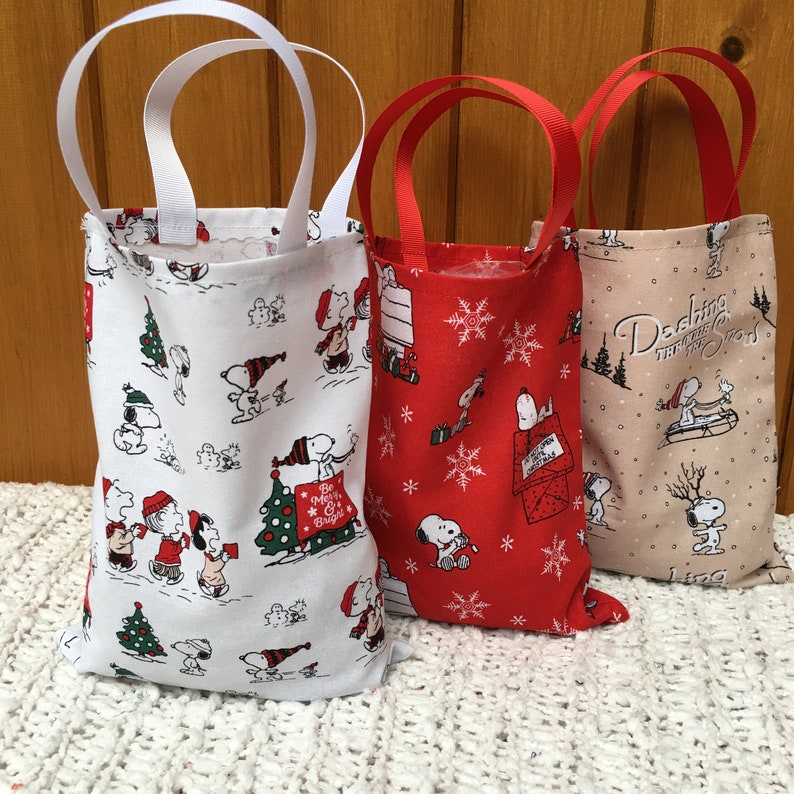 May include: Three small fabric gift bags with Christmas-themed Peanuts characters. The bags are white with red and black accents, red with white snowflakes and black accents, and beige with brown and black accents. The bags have white, red, and brown handles.