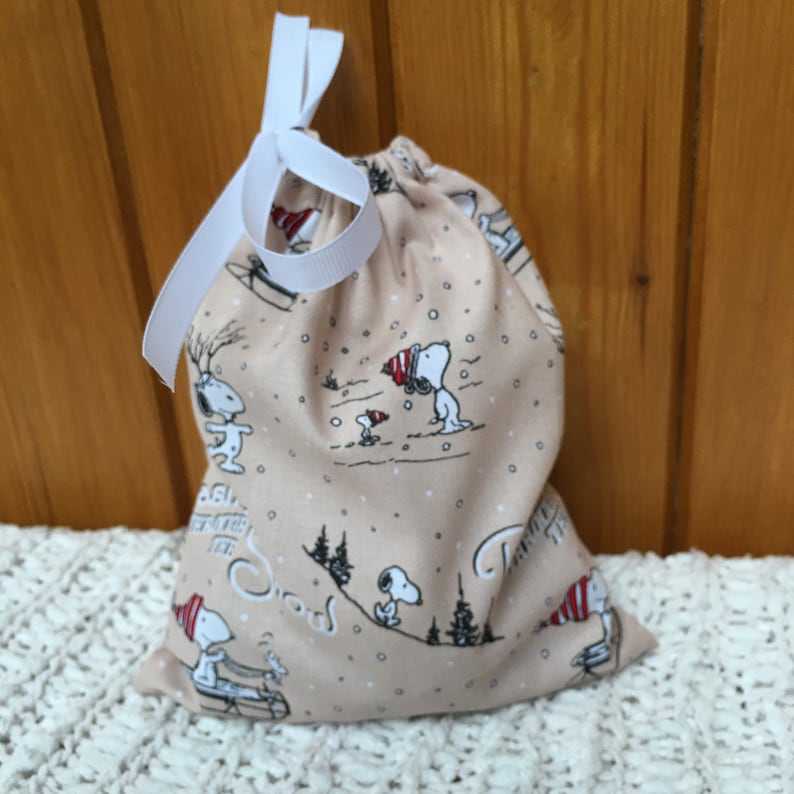 May include: A beige fabric gift bag with a white drawstring. The bag features a repeating pattern of Snoopy characters in winter scenes. The text "Snoopy" is printed on the bag.