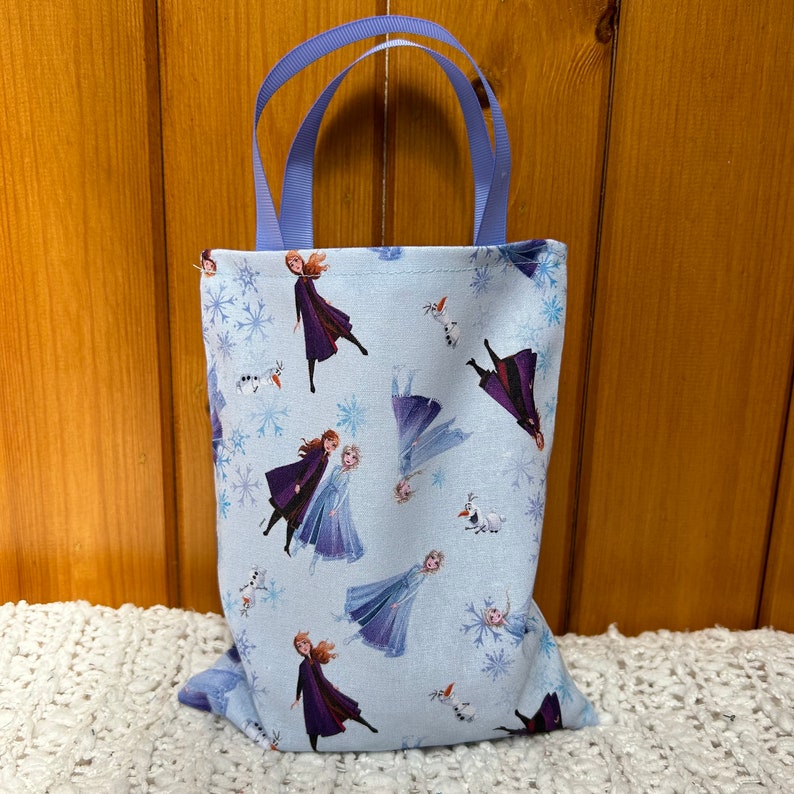 K&ouml;nnte beinhalten: Eine kleine blaue Tragetasche mit lila Henkeln. Die Tasche ist mit einem sich wiederholenden Muster der Frozen-Charaktere Elsa und Anna in verschiedenen Posen verziert. Der Hintergrund ist hellblau mit Schneeflocken.