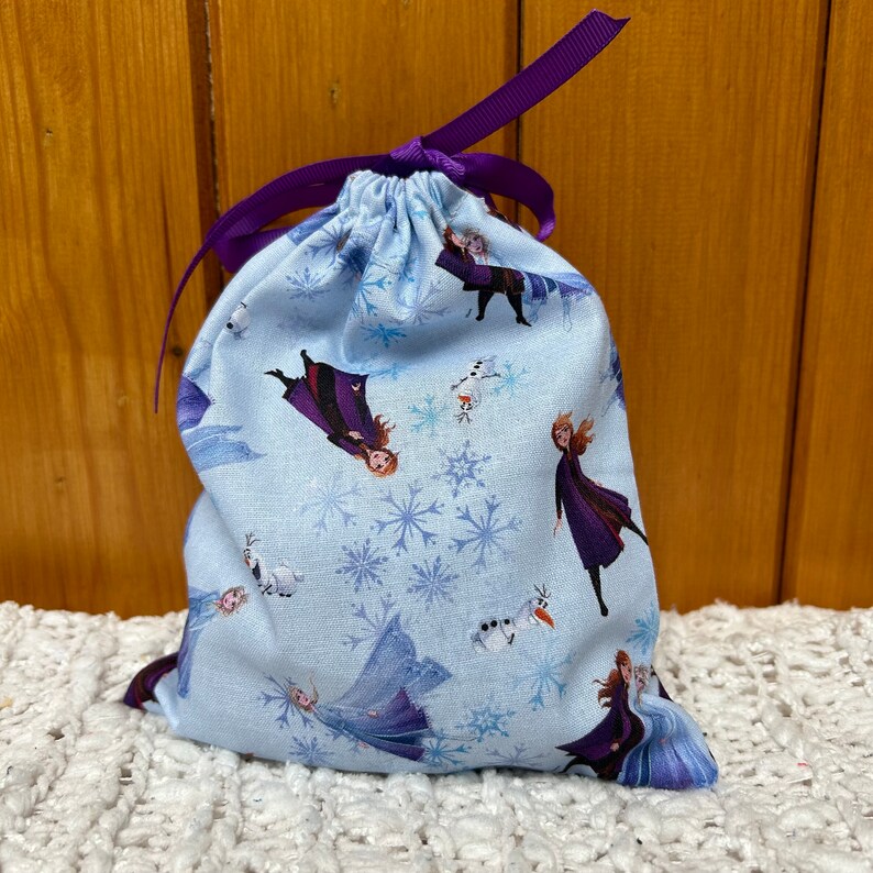 K&ouml;nnte beinhalten: Eine hellblaue Kordeltasche mit einem violetten Band. Die Tasche hat ein sich wiederholendes Muster aus Schneeflocken und Figuren aus dem Film Frozen.