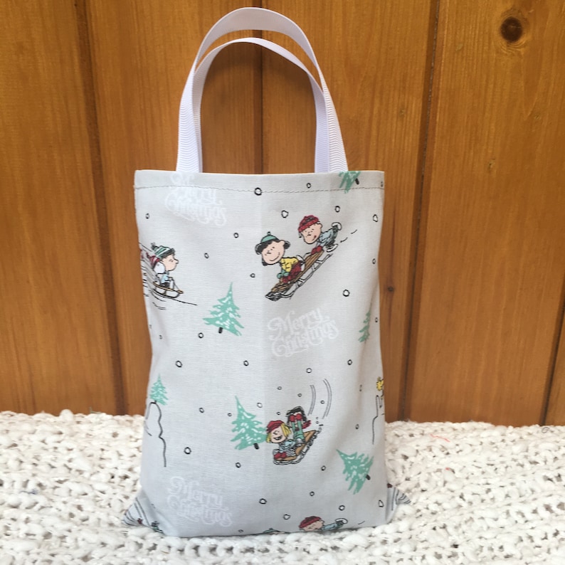 May include: A gray fabric tote bag with white handles. The bag has a repeating pattern of Peanuts characters sledding in the snow with the text "Merry Christmas".