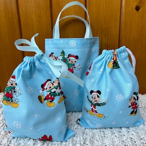 May include: Three blue Christmas gift bags with a Mickey Mouse and Minnie Mouse holiday design. The bags are made of fabric and have a drawstring closure. The bags are decorated with snowflakes and a Christmas tree.