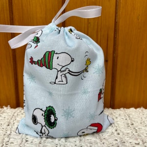 May include: A light blue drawstring bag with a white drawstring. The bag is decorated with a repeating pattern of Snoopy characters wearing Christmas hats and holding a bird. The bag is perfect for holding small gifts or treats.
