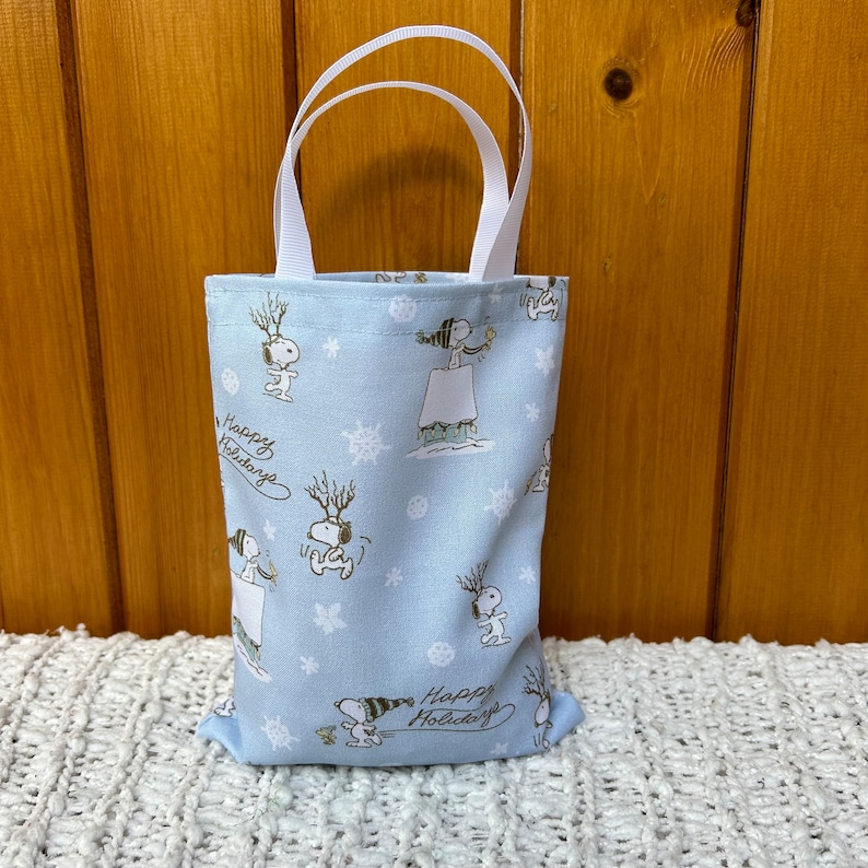 May include: A light blue reusable tote bag with white handles. The bag features a repeating pattern of Snoopy characters in winter scenes with snowflakes and the text "Happy Holidays".