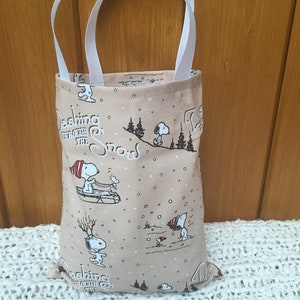 May include: A beige fabric tote bag with white handles. The bag features a repeating pattern of Snoopy characters in various winter scenes, including sledding and ice skating. The text "Dashing through the snow" is repeated on the bag.