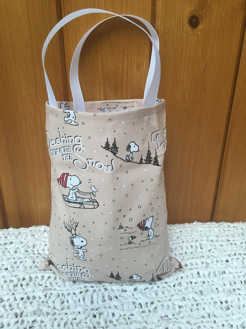 May include: A beige fabric tote bag with white handles. The bag features a repeating pattern of Snoopy characters in various winter scenes, including sledding and ice skating. The text "Dashing through the snow" is repeated on the bag.