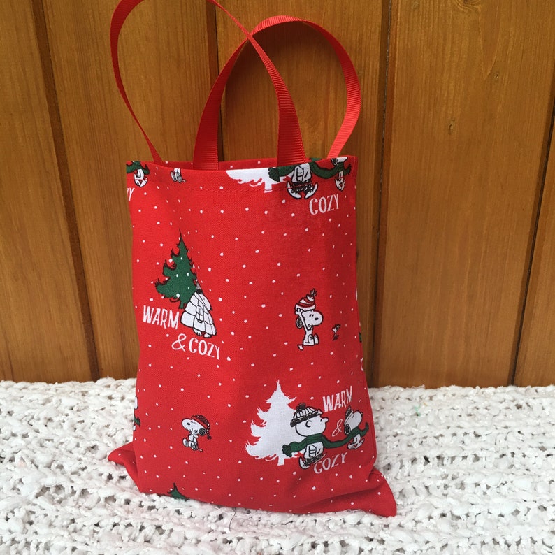 May include: Red fabric gift bag with white snowflakes and a Peanuts character design. The bag has red handles and the text "Warm & Cozy" on the front.