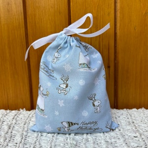 May include: A light blue drawstring bag with a white ribbon. The bag has a pattern of snowflakes and Snoopy characters. The text "Happy Holidays" is printed on the bottom of the bag.