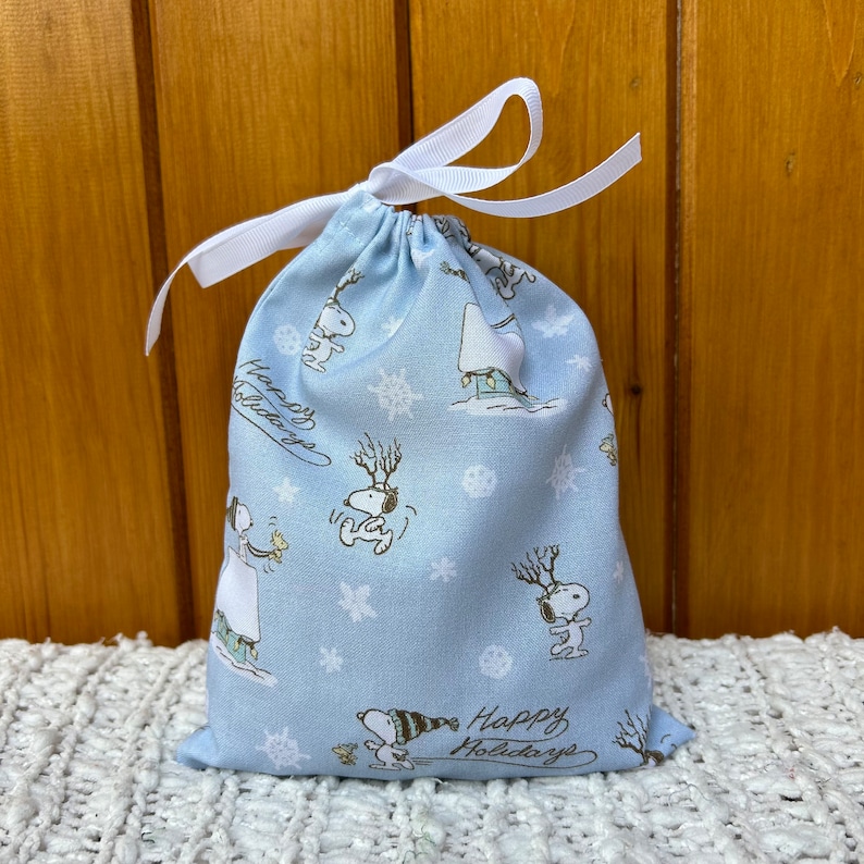 May include: A light blue drawstring bag with a white ribbon. The bag has a pattern of snowflakes and Snoopy characters. The text "Happy Holidays" is printed on the bottom of the bag.