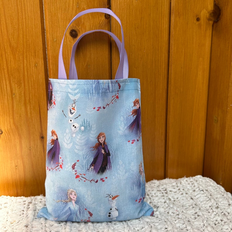 K&ouml;nnte beinhalten: Eine kleine blaue Tragetasche mit einem lilafarbenen Henkel. Die Tasche zeigt ein sich wiederholendes Muster von Figuren aus dem Film Frozen, darunter Elsa, Anna, Olaf und Kristoff.
