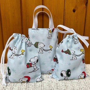 May include: Three blue fabric gift bags with a white snowflake pattern and a cartoon image of Snoopy and Woodstock. The bags are different sizes and have white drawstrings or handles.
